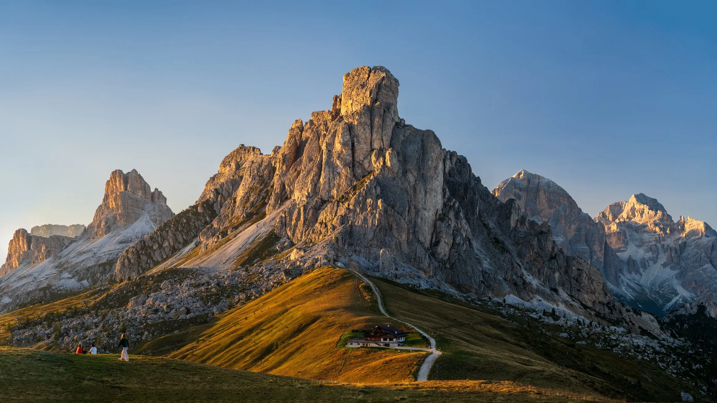 Sunlit limestone peak above a winding trail and an alpine hut.