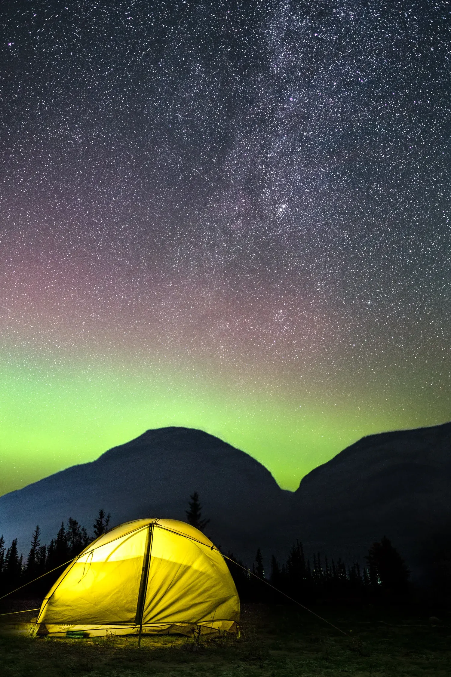 Lit tent under the Milky Way and a green aurora.